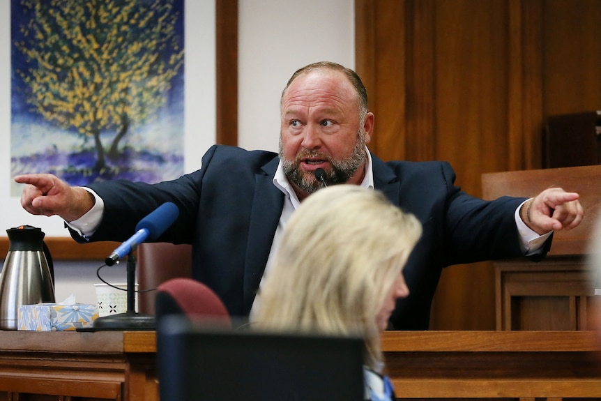 American Alex Jones sits in a courtroom witness stand, wearing a suit and pointing with both hands into the courtroom