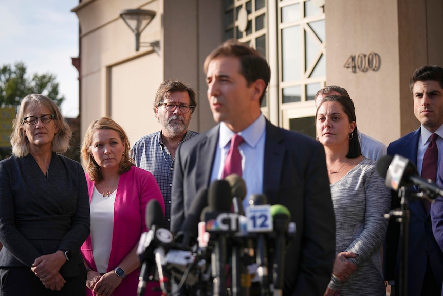 lawyer in suit speaks behind a bank of mics, three women and three men grouped behind him, looking on