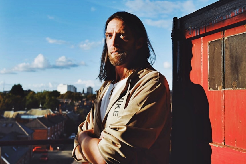 A man with shoulder-length hair stands with arms folded across chest and neutral expression, staring off to the side.