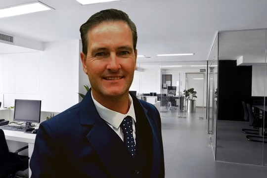 A smiling, dark-haired man in a dark suit stands in an office.