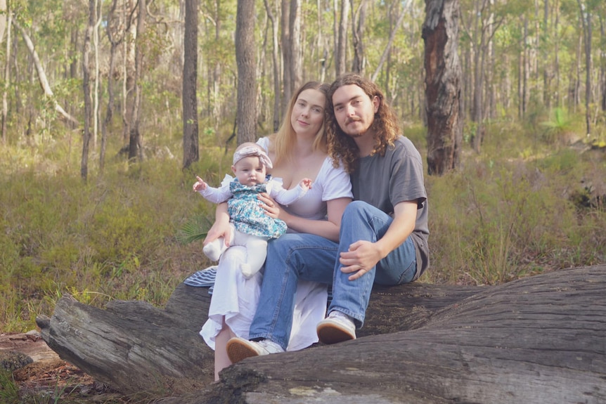 A woman with a baby on her lap leans against a man. They are sitting on a log in bushland.