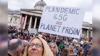 A crowd of people protest in front of a government building. In the middle, someone holds a sign reading "Plandemic & 5G = Planet Prison"