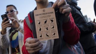 A man holds up a small piece of paper that reads "Drone Field Guide" with illustrations of different types of birds