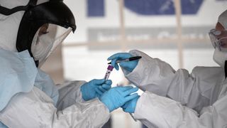 Two hospital workers in Covid PPE hand each other a test tube