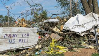 A photo of debris and wreckage from a tornado, with a painted sign reading "pray for us all"