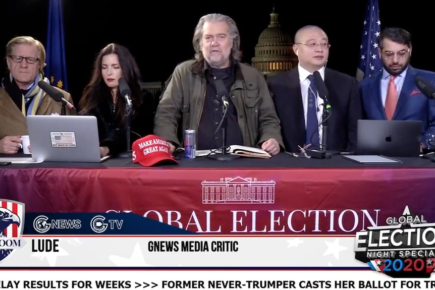 Five people sit behind a panel table presenting a broadcast with the US Capitol Building in the background