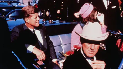 Getty Images Texas Governor John Connally adjusts his tie (foreground) as US President John F Kennedy (left) & First Lady Jacqueline Kennedy (in pink) settled in rear seats, prepared for motorcade into city from airport, Nov. 22. After a few speaking stops, the President was assassinated in the same car.