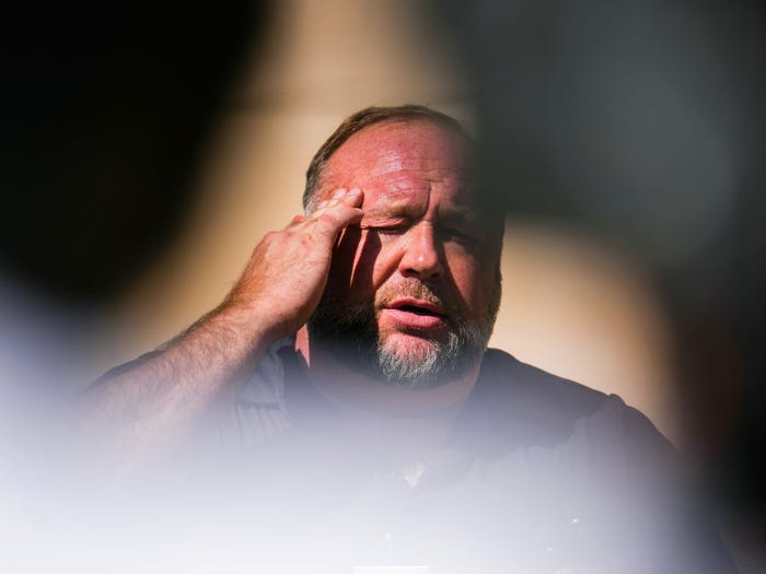 Jones outside of court in Connecticut in 2022, holding a hand to his head with his eyes closed, and blurry figures in the foreground.