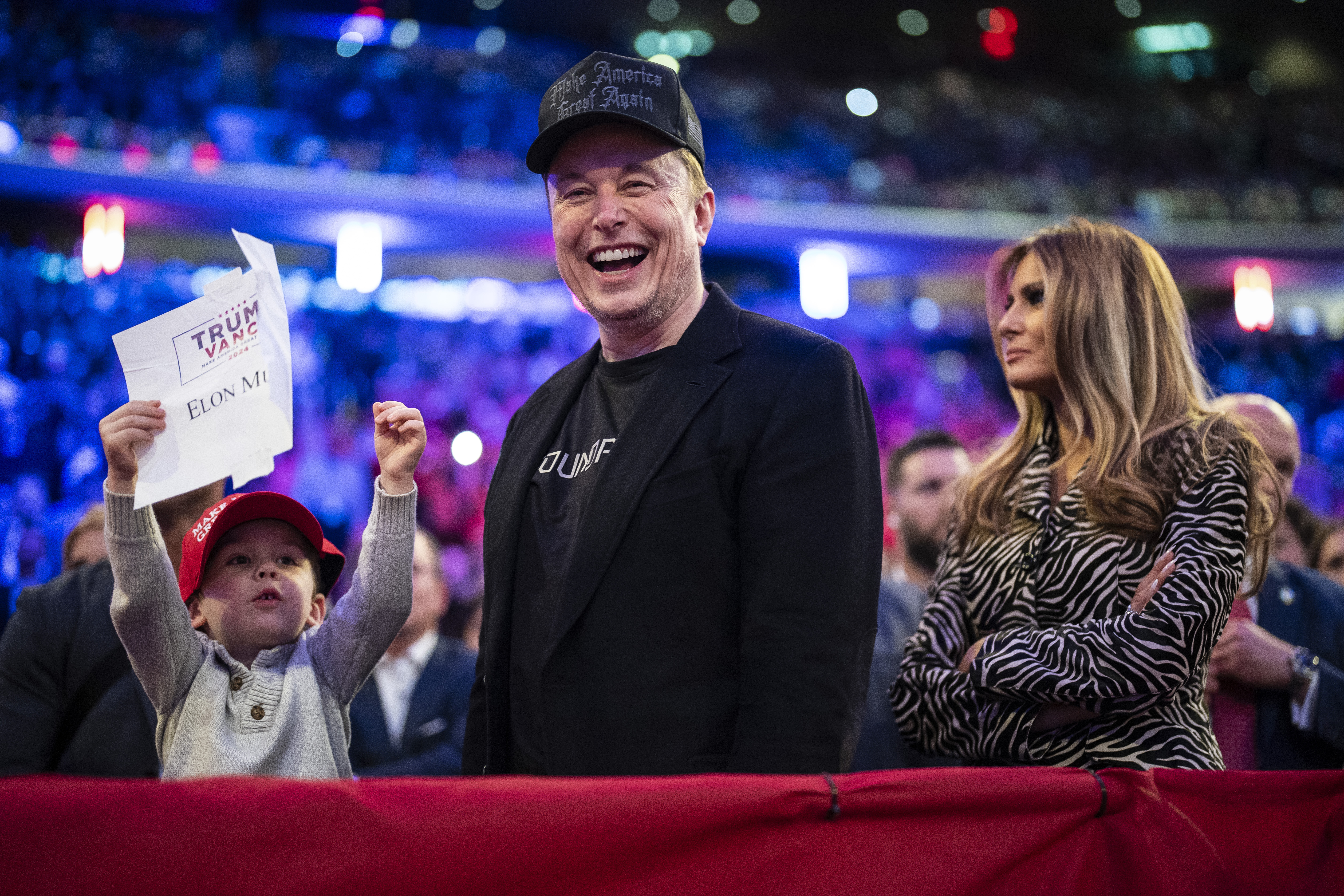 Elon Musk and Melania Trump stand with Musk’s young son at a Trump rally. The kid is waving a Trump-Vance sign.